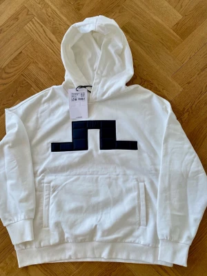 Vit hoodie från J.Lindeberg - Snygg vit hoodie från J.Lindeberg med stor svart logga på bröstet. Hoodien har huva med dragsko, magficka och ribbade muddar vid ärmar och nederkant. Tillverkad i mjukt material som känns skönt mot huden. Perfekt för en avslappnad och trendig look.