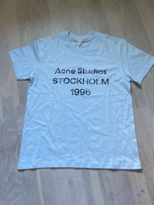 Vit Acne Studios t-shirt Stockholm - Vit t-shirt från Acne Studios med svart text 'Acne Studios STOCKHOLM 1996' på bröstet. Klassisk rund hals och korta ärmar. Perfekt basic-plagg med stilren design och skön passform.