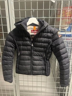Svart dunjacka från Parajumpers - Snygg svart dunjacka från Parajumpers med huva och dragkedja framtill. Jackan har två sidofickor med dragkedja och ett tydligt Parajumpers-märke på ärmen. Lätt vadderad och perfekt för kalla vinterdagar. ( det är en barn jacka den passar för 11 åringar och neråt )