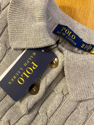 Grå kabelstickad polotopp från Polo Ralph Lauren - Snygg grå kabelstickad polotopp från Polo Ralph Lauren med korta ärmar och klassisk krage. Toppen har knappdetalj framtill och den ikoniska loggan broderad på bröstet. Perfekt till jeans eller kjol för en clean och stilren look.