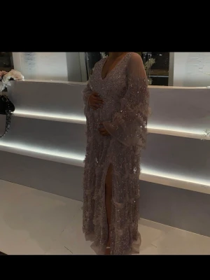 SÖKER‼️‼️Rosa glittrig maxiklänning med slits  - Denna klänning sökes storlek 40/42‼️.  Asos Elegant rosa maxiklänning med glittriga paljetter, djup v-ringning och lång slits framtill. Klänningen har vida, transparenta ärmar och dekorerade volangdetaljer som ger en lyxig känsla. Perfekt för speciella tillfällen.