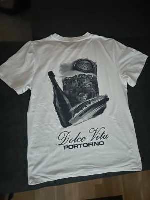 Vit Dolce Vita Portofino t-shirt - Säljer en vit t-shirt med tryck både fram och bak. På ryggen finns ett stort svartvitt motiv med texten 'Dolce Vita Portofino', en flaska, båt och stadsmotiv. Framtill står samma text i mindre format. Klassisk rund hals och korta ärmar.