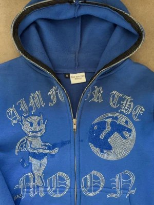Aim for the moon hoodie - Blå zip-hoodie med huva och coolt motiv framtill i strasstenar. Texten 'AIM FOR THE MOON' och en tecknad figur med vingar och gitarr ger en unik streetwear-vibe. Mjuk insida och ribbade muddar. Perfekt för dig som gillar statement-plagg.