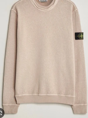 Beige sweatshirt från Stone Island - Snygg beige sweatshirt från Stone Island med klassisk rund hals och ribbade muddar. Tröjan har den ikoniska loggan på vänster ärm och är tillverkad i mjukt material som känns skönt mot huden. Perfekt för en avslappnad och stilren look.