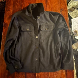 Svart mocka overshirt med fickor - Stilren svart mocka overshirt med knäppning framtill och två bröstfickor med lock och knappar. Rak passform och klassisk krage. Perfekt lager-på-lager-plagg för en avslappnad look. En knapp saknas 