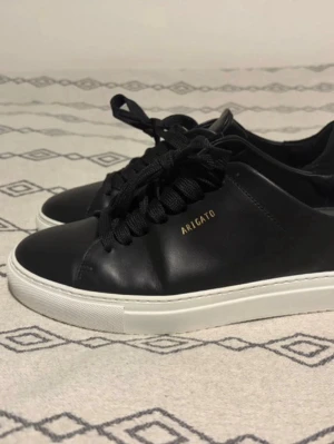 Svarta sneakers från Axel Arigato - Stilrena svarta sneakers från Axel Arigato med vit sula och guldfärgad logga på sidan. Skorna har ovandel i slätt läder, svarta skosnören och detaljer i mocka vid hälen. Perfekta för en clean och modern look.