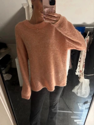 Ljusrosa fluffig stickad tröja - Mysig ljusrosa stickad tröja med rund hals och lång ärm. Tröjan har en fluffig, mjuk struktur och avslappnad passform. Perfekt för kyliga dagar och enkel att styla med jeans eller kjol.