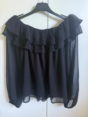 Svart offshoulder blus med volanger - Trendig svart offshoulder blus med dubbla volangdetaljer runt ringningen. Tillverkad i ett tunt, transparent tyg som ger en luftig känsla. Långa ärmar med lätt puff och elastiska muddar. 