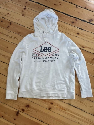 Lee hoodie med tryck - Snygg vit hoodie från Lee med tryck framtill i blått och rött där det står 'Lee', 'Salina Kansas' och 'Lee Denim'. Klassisk huva med dragsko och långa ärmar. Perfekt för en avslappnad och cool stil.