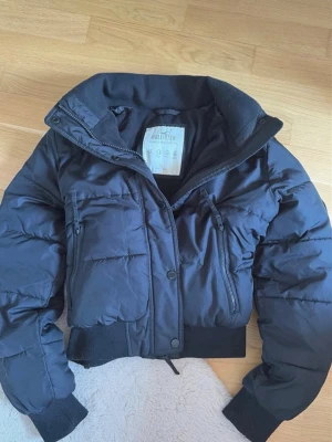 Svart pufferjacka från Hollister XS - Snygg svart pufferjacka från Hollister Ultimate Puffer Collection. Jackan har hög krage, två stora bröstfickor med dragkedja och ribbade muddar vid ärmar och midja. Perfekt för kalla dagar 