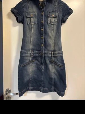 Kort jeansklänning med knappar - Snygg kort jeansklänning i blå denim med tryckknappar framtill, två bröstfickor och korta ärmar. Klänningen har markerad midja och dragkedjedetaljer vid fickorna. Perfekt för en avslappnad och trendig look.