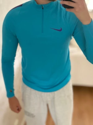 Blå långärmad träningströja Nike - Snygg blå långärmad träningströja från Nike med halv dragkedja framtill och lila detaljer på axlarna samt lila Nike-logga på bröstet. Tillverkad i lätt och funktionellt material med Dri-FIT-teknologi som transporterar bort fukt. Perfekt för träning eller uppvärmning.