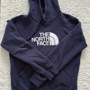 Mörkblå hoodie från The North Face - Snygg mörkblå hoodie från The North Face med stor vit logga på bröstet. Klassisk känguruficka framtill, justerbar huva med dragsko och ribbade muddar vid ärmar och midja. Perfekt för en avslappnad och trendig stil.