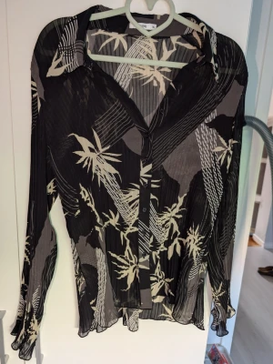 Mönstrad, plisserad blus - Snygg svart blus med beige och vita bambuliknande mönster. Blusen har knappar framtill, krage och är tillverkad i ett tunt, plisserat material som ger en luftig känsla. Långärmad modell med avslappnad passform, perfekt för en trendig look.