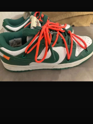 Nike Dunk Low Off-White Pine green - Nike Dunk Low Off-White sneakers i grönt och vitt med dubbla snörningar, där de yttre snörena är orange. Skon har klassisk låg siluett, perforerad tåbox och detaljerad Off-White design. Ovandelen är i läder och mesh för en sportig vibe.