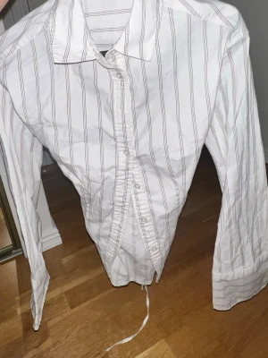 Vit randig skjorta med snörning - Säljer en vit skjorta med tunna grå ränder och lång ärm. Skjortan har knappar framtill och snörning i ryggen för en snygg detalj. Perfekt för dig som gillar en twist på den klassiska skjortan. Man kan justera den där bak och göra så den blir extra tajt om man vill det eller så kan man ha den ganska lös båda sätten är jätte fina💕