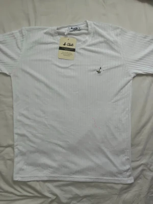 Vit ribbad t-shirt från Club - Snygg vit t-shirt från Club med diskret ribbad struktur och liten broderad fågel på bröstet. Klassisk rund halsringning och korta ärmar. Perfekt basplagg för en clean och stilren look.