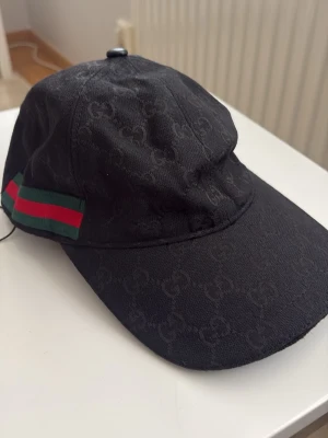Svart keps  - Snygg svart keps från med diskret monogram över hela tyget och klassiska gröna och röda ränder på sidan. Justerbar rem baktill i läder och böjd skärm. Perfekt accessoar för en trendig och exklusiv look.