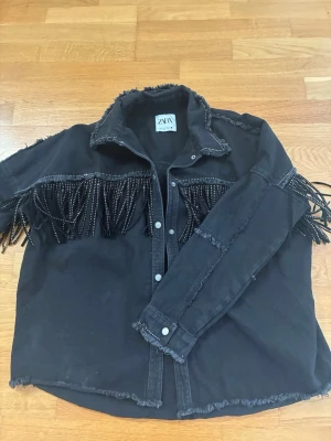 Svart jeansjacka med fransar från Zara - Cool svart jeansjacka från Zara med råa kanter och dekorativa fransar med pärldetaljer över bröstet och armarna. Jackan har tryckknappar framtill och en avslappnad passform. Perfekt för dig som vill sticka ut med en unik och trendig look.