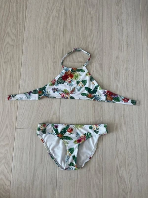 Vit bikini med blommigt mönster - Snygg bikini i vitt med färgglatt blommigt mönster och gröna blad. Underdelen har klassisk bikini-modell. Bikiniöverdelen har som ett knäppe på ryggen. Perfekt för strandhäng och sommarens alla bad.