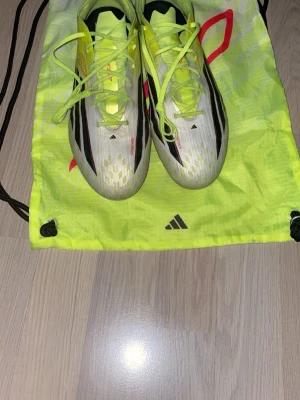 Adidas gula och vita fotbollsskor - Säljer ett par snabba fotbollsskor från Adidas i vitt och neongult med svarta detaljer. Skorna har klassisk snörning och en lätt, strömlinjeformad design som passar perfekt för snabba spelare. Lite tuggummi bit till vänster om vänster skon. Medföljer en matchande neongul påse med Adidas-logga. 