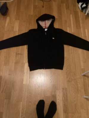 Svart zip hoodie från Burberry - Säljer en svart hoodie från Burberry med dragkedja och huva. Insidan av huvan har det klassiska Burberry-mönstret med beige och röd detalj. Liten Burberry-logga broderad på bröstet. Perfekt för dig som vill ha en stilren och exklusiv look.