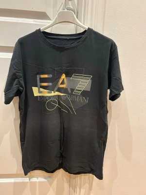 Svart EA7 t-shirt med gulddetaljer - Snygg svart t-shirt från Emporio Armani EA7 med stort tryck i guld och gult framtill. Klassisk rund halsringning och korta ärmar. Perfekt för dig som gillar stilrena plagg med en touch av lyx.