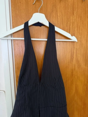 Fest topp  - Snygg svart fest topp  med vita smala kritstrecksränder och djup v-ringning. Halterneck-modell som går runt nacken och har en figurnära passform upptill. Perfekt för dig som gillar en trendig och stilren look.