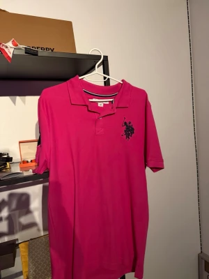 Rosa pikétröja med tryck - Snygg rosa pikétröja med krage och korta ärmar. Har ett stort tryck på bröstet och siffror på ena ärmen. Perfekt för dig som vill sticka ut med färg. Tillverkad i bomull för skön känsla.