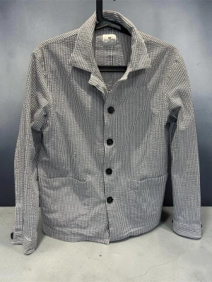 Randig overshirt från John Henric  - Snygg overshirt i blåvitt randigt tyg med fyra stora svarta knappar framtill från John henric . Två rymliga fickor framtill och klassisk krage. Långärmad modell med knapp vid ärmslut. Perfekt lager-på-lager-plagg för en avslappnad stil.