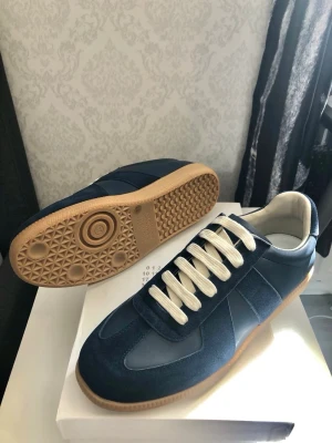 Maison Margiela - Snygga mörkblå sneakers med beige gummisula och vita snören. Ovandelen är i en mix av mocka och skinn för en stilren look. Perfekta för dig som vill ha klassiska men trendiga skor till vardags. Levereras i originalkartong.
