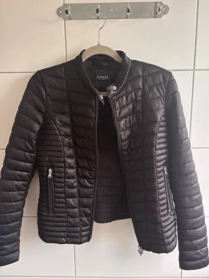 Svart pufferjacka från Guess - Snygg svart pufferjacka från Guess med quiltad design och raka linjer. Jackan har ståkrage med tryckknapp, dragkedja framtill och två fickor med dragkedja. Materialet är glansigt och ger en modern look. Perfekt för kalla dagar.