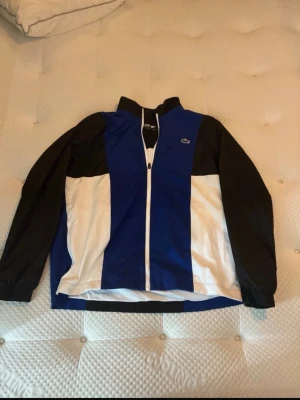 Blå och svart vindjacka och byxor  från Lacoste - Snygg vindjacka från Lacoste i blått, svart och vitt med dragkedja framtill och hög krage. Jackan har långa ärmar och logga på bröstet samt stor Lacoste-text och krokodil på ryggen. Tillverkad i lätt och tunt material, perfekt för blåsiga dagar.