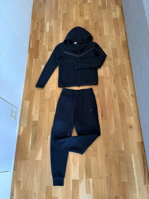 Svart Nike Tech Fleece Set - Säljer ett komplett Nike Tech Fleece set i färgen svart. Både jackan (tröjan med dragkedja) och byxorna är i storlek S. Hela setet är köpt nytt under 2023.  Tröjan är inköpt på Zalando (nypris: 1281 kr).  Byxorna är inköpta direkt från Nikes hemsida (nypris: 987 kr). Det är knappt använt och i ett jättefint skick, precis som bilderna visar. Det har alltid tvättats varsamt och har inga synliga slitage eller fläckar. Självklart är det helt äkta. Jag har kvar orderbekräftelserna digitalt.