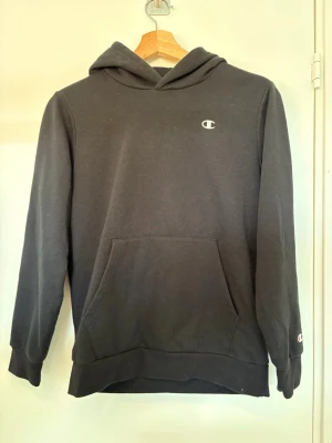 Svart hoodie från Champion - Svart hoodie från Champion med klassisk logga på bröstet och ärmen. Hoodie är en lite tunnare modell, i storleken ( cm 150/155, och åldern 11/12). 