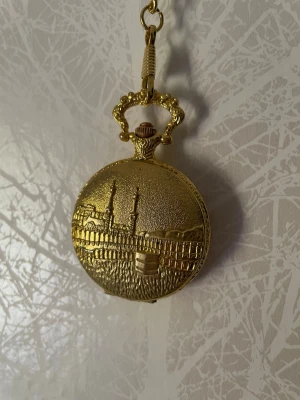 Medina/Mecka Fickur från Saudiarabien - Säljer ett guldfärgat fickur med lång kedja, och ett detaljerat motiv på locket som föreställer de heliga städerna Medina på ena sidan och Mecka på andra sidan. Uret har tyvärr gått ur funktion och behöver fixas. Annars funkar den utmärkt som en dekorativ accessoar.   OBS: En liten del av guldfärgen på Mecka-sidan har slitits bort. Detta är en ursprunglig defekt.