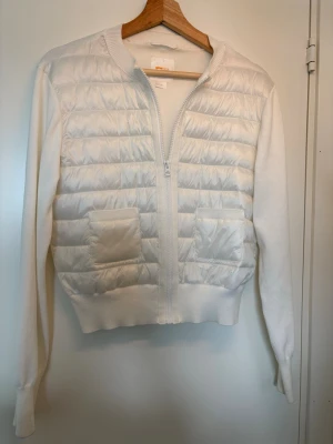 Vit quiltad cardiganjacka - Stilren vit cardiganjacka med quiltad framsida och två fickor. Jackan har dragkedja, ribbade muddar och är tillverkad i en mix av stickat tyg och vadderat material framtill. I storleken 170.
