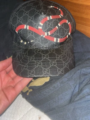 Gucci keps med orm och GG-mönster - Snygg svart keps från Gucci med klassiskt GG-mönster och en röd orm på framsidan. Justerbar rem bak med blå och röd rand. Kepsen har mesh på baksidan för extra ventilation och är tillverkad i slitstarkt material.