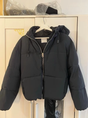 Svart pufferjacka med huva - Säljer en svart pufferjacka med dragkedja framtill och stor huva. Jackan har en boxig passform, dragsko i huvan och är tillverkad i ett vadderat material som håller värmen. Perfekt för kalla vinterdagar och riktigt skön att bära.