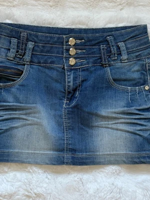 Blå jeanskjol med knappar - Snygg blå jeanskjol i kort modell med tre dekorativa silverfärgade knappar framtill och klassiska fickor. Kjolen har bred linning med bälteshällor och dragkedja vid fickan. Perfekt för en trendig och avslappnad stil.