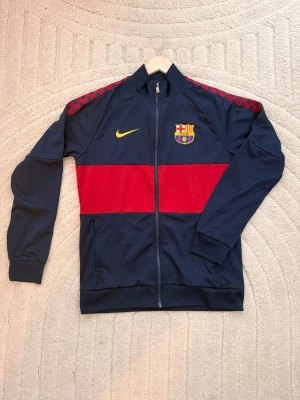 Blå Barcelona träningsjacka Nike - Snygg blå träningsjacka från Nike med FC Barcelona-logga på bröstet och gul Nike-swoosh. Jackan har en röd rand över bröstet och rutiga detaljer på axlarna. Hel dragkedja framtill och ståkrage. Perfekt för fotbollsträning eller som sportig vardagsstil.