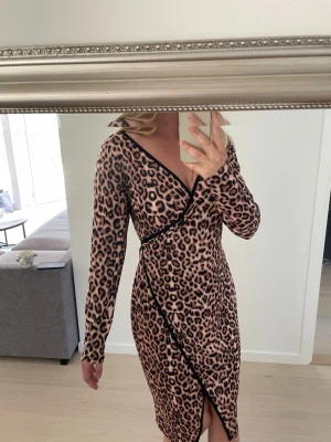 Leopardmönstrad omlottklänning - Säljer en leopardmönstrad omlottklänning i beige och svart med lång ärm och v-ringning. Klänningen har ett knytband i midjan och slits framtill för en snygg siluett. Passar storlek S till L då det är justerbara band i midjan. 