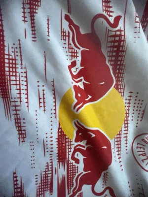 RB Leipzig vit röd matchtröja - Säljer en vit matchtröja med rött och gult tryck från RB Leipzig. Tröjan har två röda tjurar mot en gul cirkel på bröstet och röda grafiska detaljer. Tillverkad i ett lätt och snabbtorkande material, perfekt för träning eller match.Redbull märke, size 134, 8-9 år