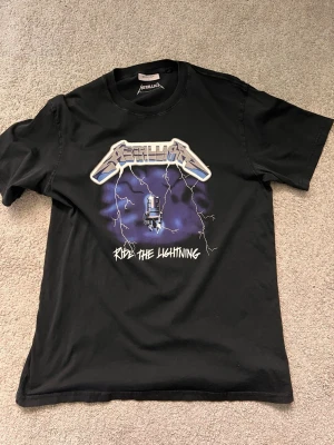 Svart Metallica Ride the Lightning t-shirt - Cool svart t-shirt med Metallica Ride the Lightning-tryck på bröstet. Stor logga och motiv i blått och vitt med blixtar och elstolpe. Rund halsringning och korta ärmar. Perfekt för dig som gillar bandmerch och rockstil.