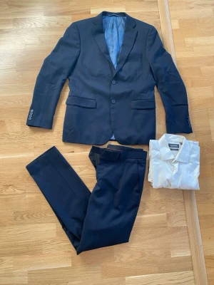 Mörkblå kostym med vit skjorta - Snygg mörkblå kostym med tvåknäppt kavaj och matchande byxor från dressman. Kavajen har klassisk passform, fickor med lock och knappar vid ärmslut. Medföljer en vit skjorta med krage.   Kavaj: strl 48 regular Byxa: strl 32/34 Skjorta: strl M