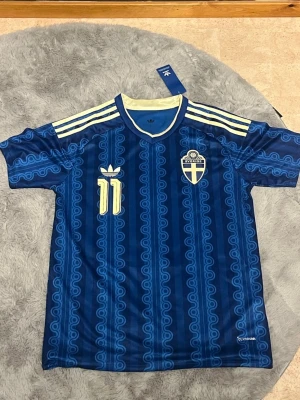 Blå Adidas fotbollströja Elanga - Snygg blå fotbollströja från Adidas med Elanga och nummer 11 på ryggen. Tröjan har ett unikt mönster i olika blå nyanser, gula detaljer och klassiska Adidas-ränder vid kragen. Perfekt för dig som gillar fotboll och vill sticka ut på planen.
