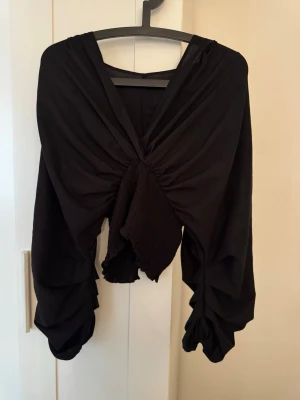 Svart crop top med draperade ärmar - Trendig svart crop top med djup v-ringning och draperade, vida ärmar. Toppen har en twistad detalj framtill och är gjord i ett mjukt, tunt tyg som ger en cool och edgy look. Perfekt för dig som vill sticka ut med din stil.