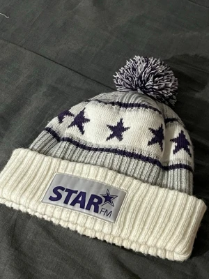 Star fm mössa - Stickad mössa i vitt och ljusgrått med mörklila stjärnmönster och en stor boll på toppen. Upprullad kant med ett tygmärke där det står STAR FM. Perfekt för kalla dagar och riktigt mysig stil.