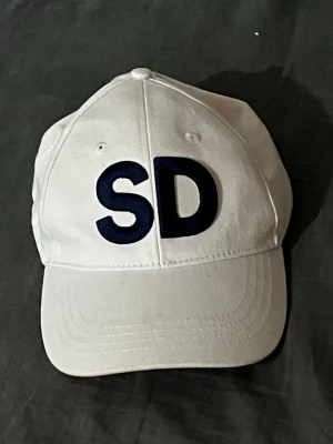 SD keps  - Snygg vit keps med böjd skärm och stora svarta bokstäver 'SD' broderade framtill. Klassisk modell med justerbar rem bak och ventilationshål på ovansidan. Perfekt accessoar för en avslappnad streetwear-look.