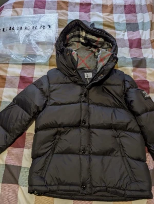 Svart dunjacka med huva från Burberry - Snygg svart dunjacka från Burberry med avtagbar huva och klassiskt rutigt foder. Jackan har dragkedja och knappar framtill, två sidofickor med dragkedja och en patch på ärmen. Perfekt för kalla vinterdagar och riktigt varm.